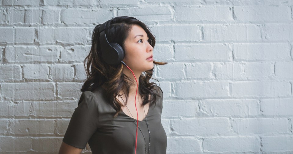 A woman standing against a white brick wall listening through headphones