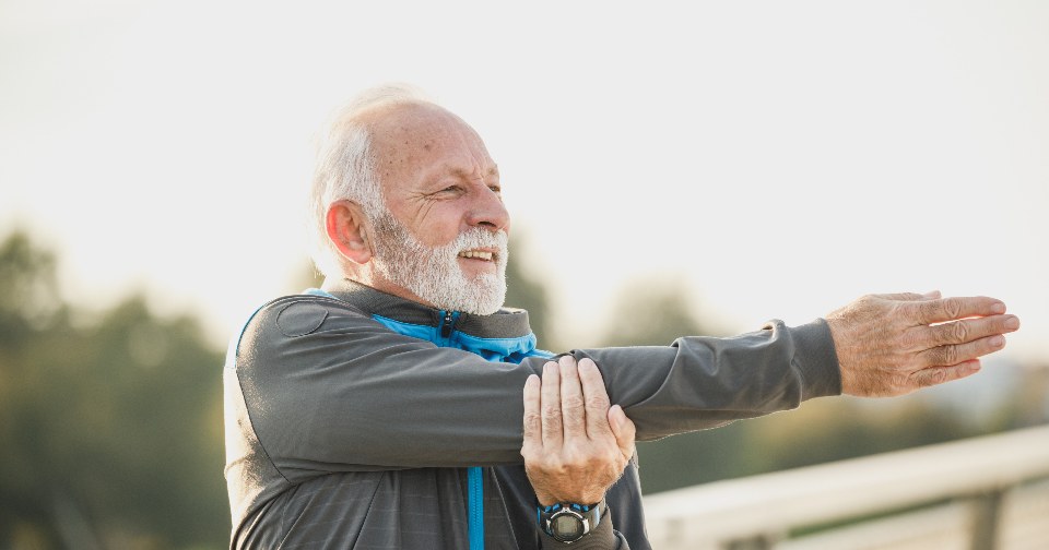 Older man in gray jacket stretching arms by a waterfront on a calm morning